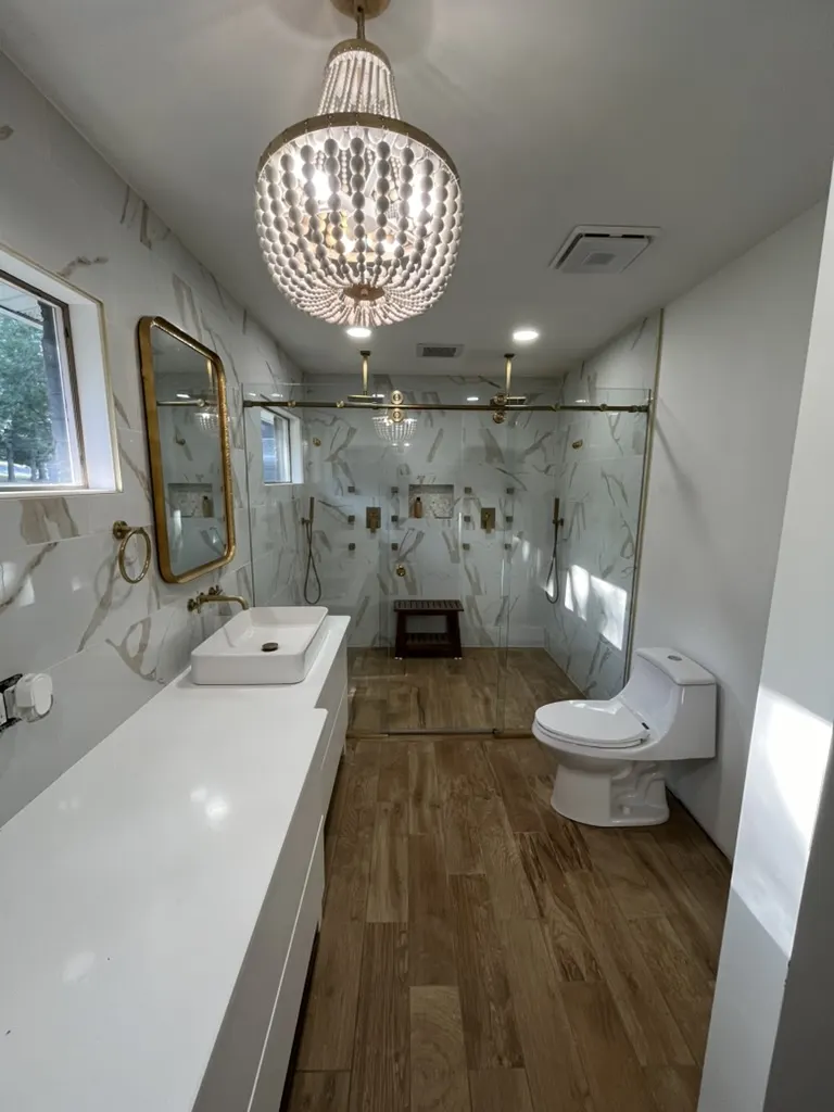 Walk-in glass shower panel in a narrow marble-look bathroom with a beaded chandelier and brass fixtures.