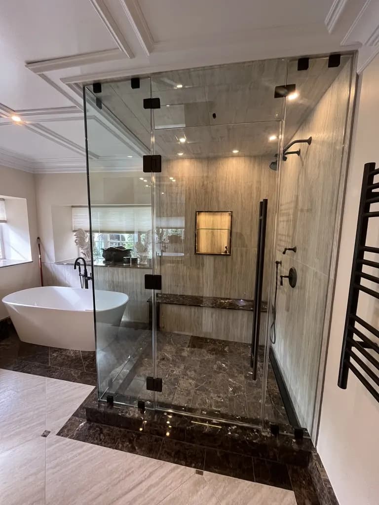 Large frameless steam enclosure in travertine-look tile with a dark marble bench and floor, beside a freestanding tub.
