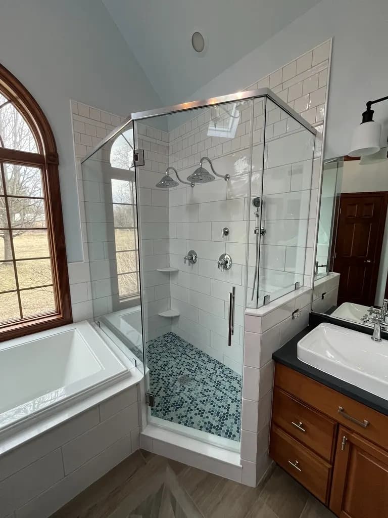 Neo-angle semi-frameless shower enclosure with twin shower heads beside a soaking tub and an arched window.