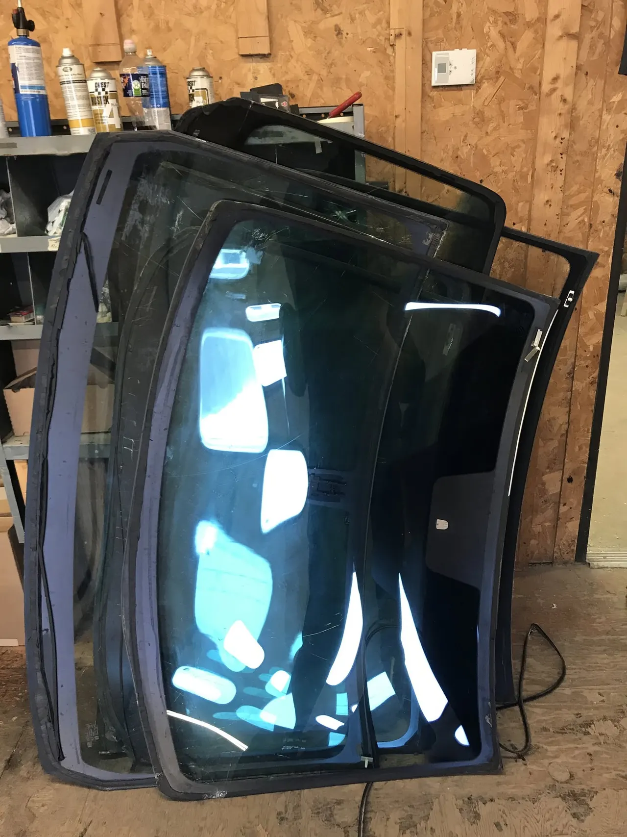 Vehicle back glass panels leaning against a plywood shop wall beside adhesive and primer cans