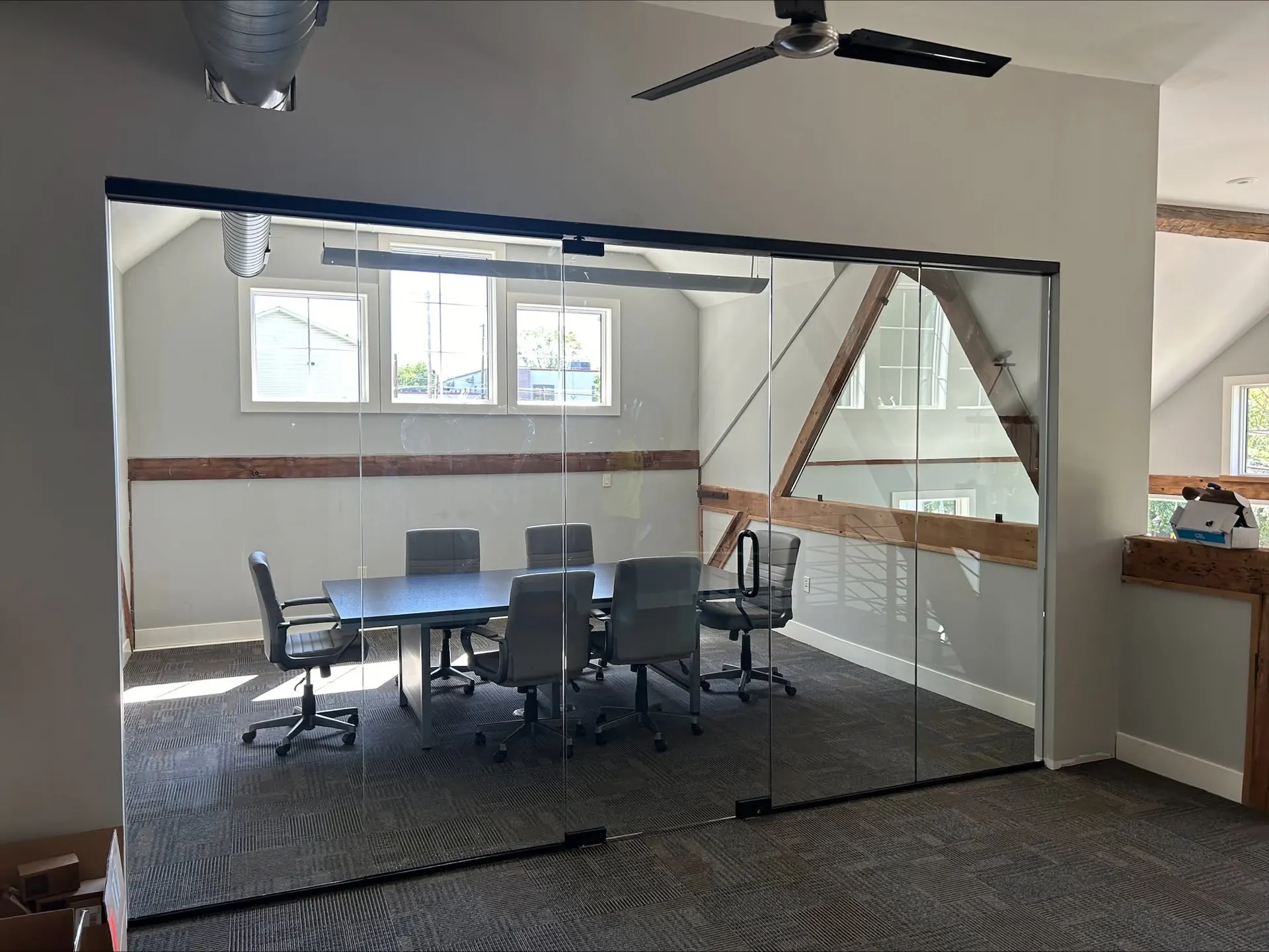 Glass-walled conference room with a long table and six chairs, exposed roof trusses and windows beyond the glass.