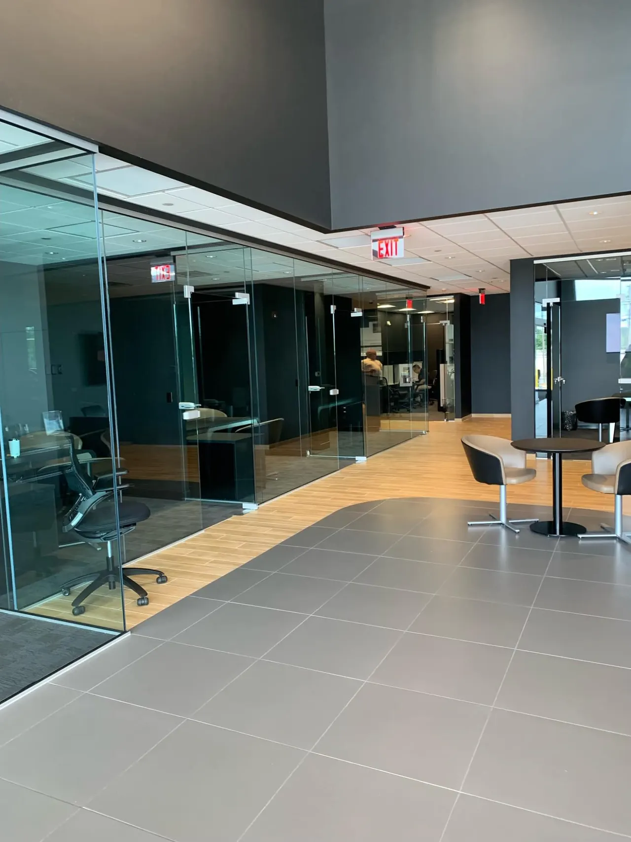 Commercial office corridor with frameless glass partition walls, black tile flooring and a small lounge seating area.