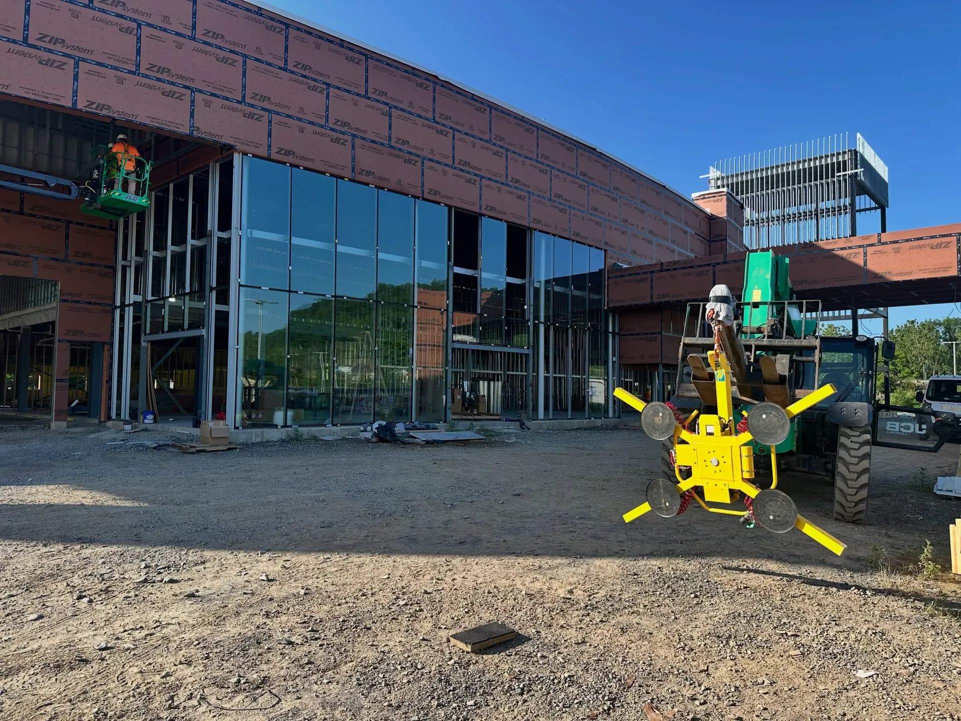 Two-story glass curtain-wall storefront under construction. Crew on a scissor lift, glass vacuum lifter staging a panel.