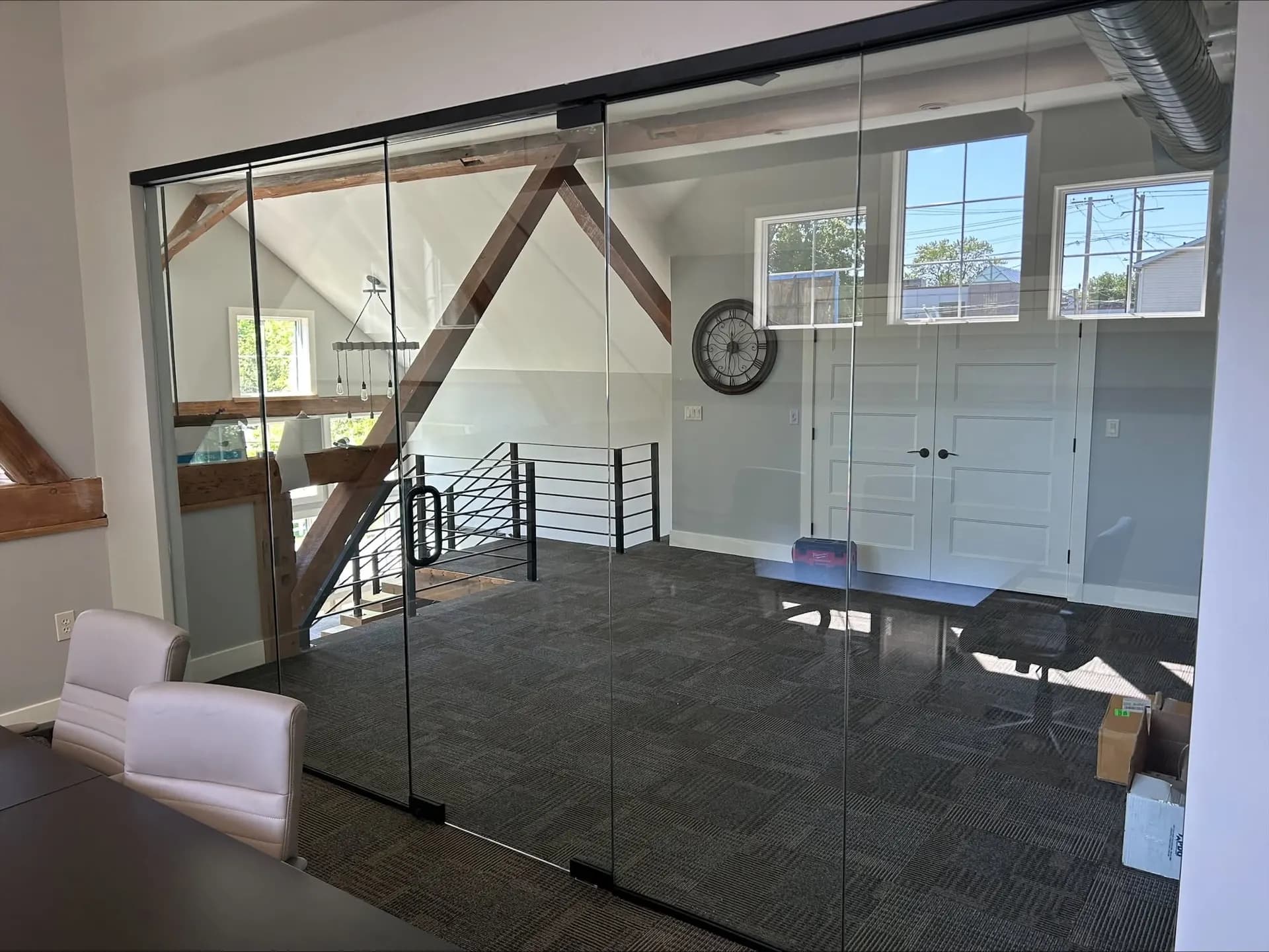 Frameless glass sliding doors dividing an open loft office, exposed wood roof trusses and a wall clock visible beyond.