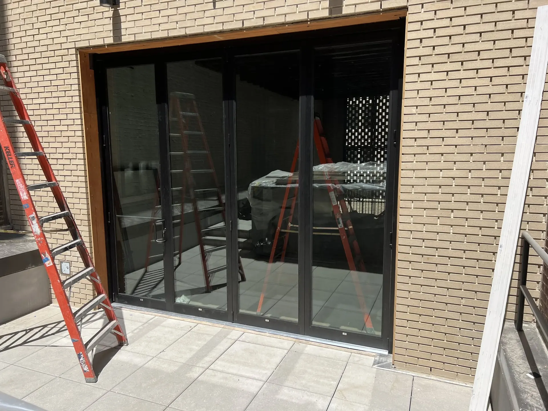 Wide black-framed sliding glass door system set into a tan brick storefront wall above a tiled patio