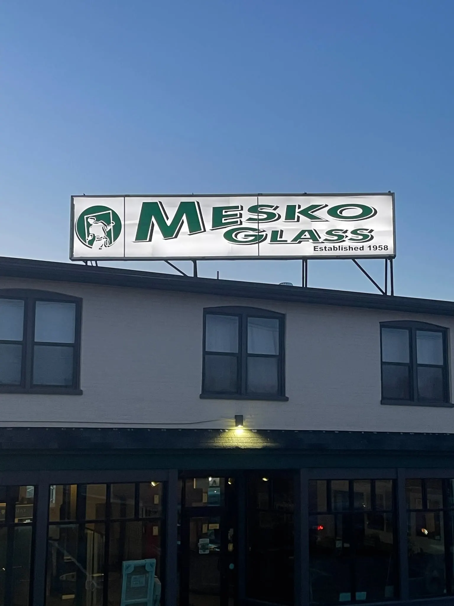Illuminated Mesko Glass rooftop sign at dusk, reading Established 1958, above the East Stroudsburg showroom.