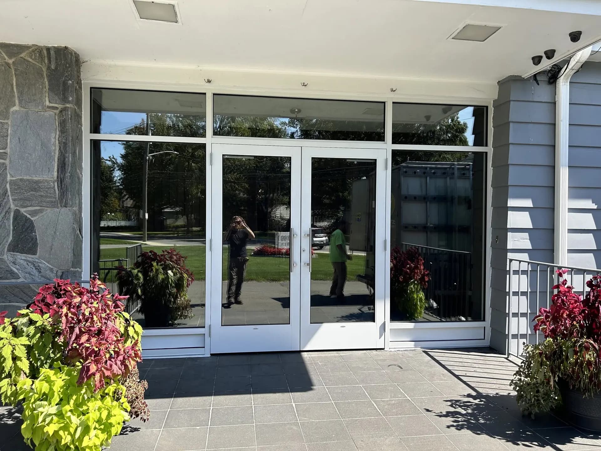 Aluminum-framed commercial entrance with double glass doors and glass sidelights, flanked by flower planters.
