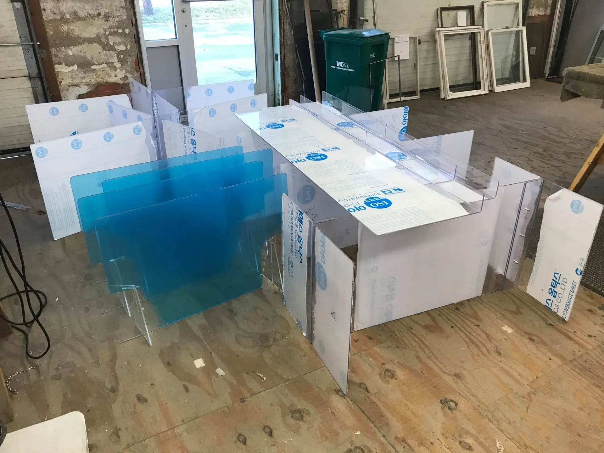 Acrylic barrier panels and film-covered sheet stock standing on the shop floor beside old sashes.