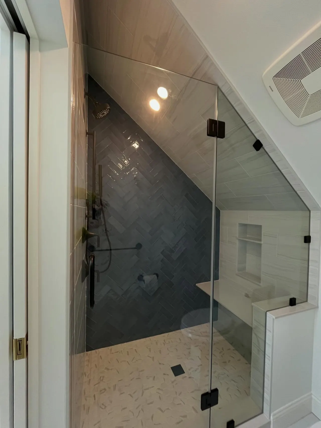 Frameless glass shower enclosure with black hardware, built into a sloped ceiling nook with herringbone tile.