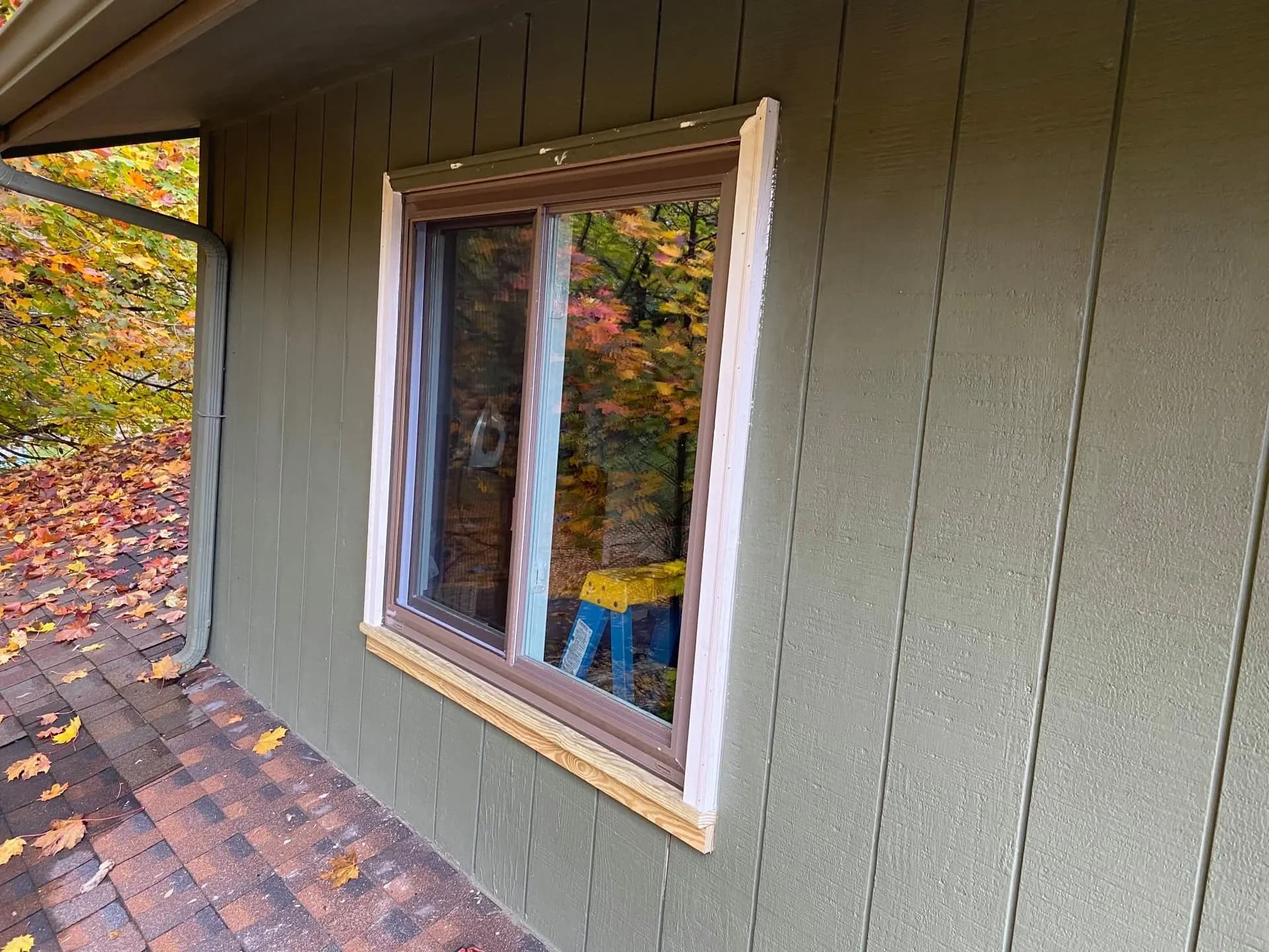 New white slider window in fresh wood trim on an olive board-and-batten wall, autumn leaves outside.