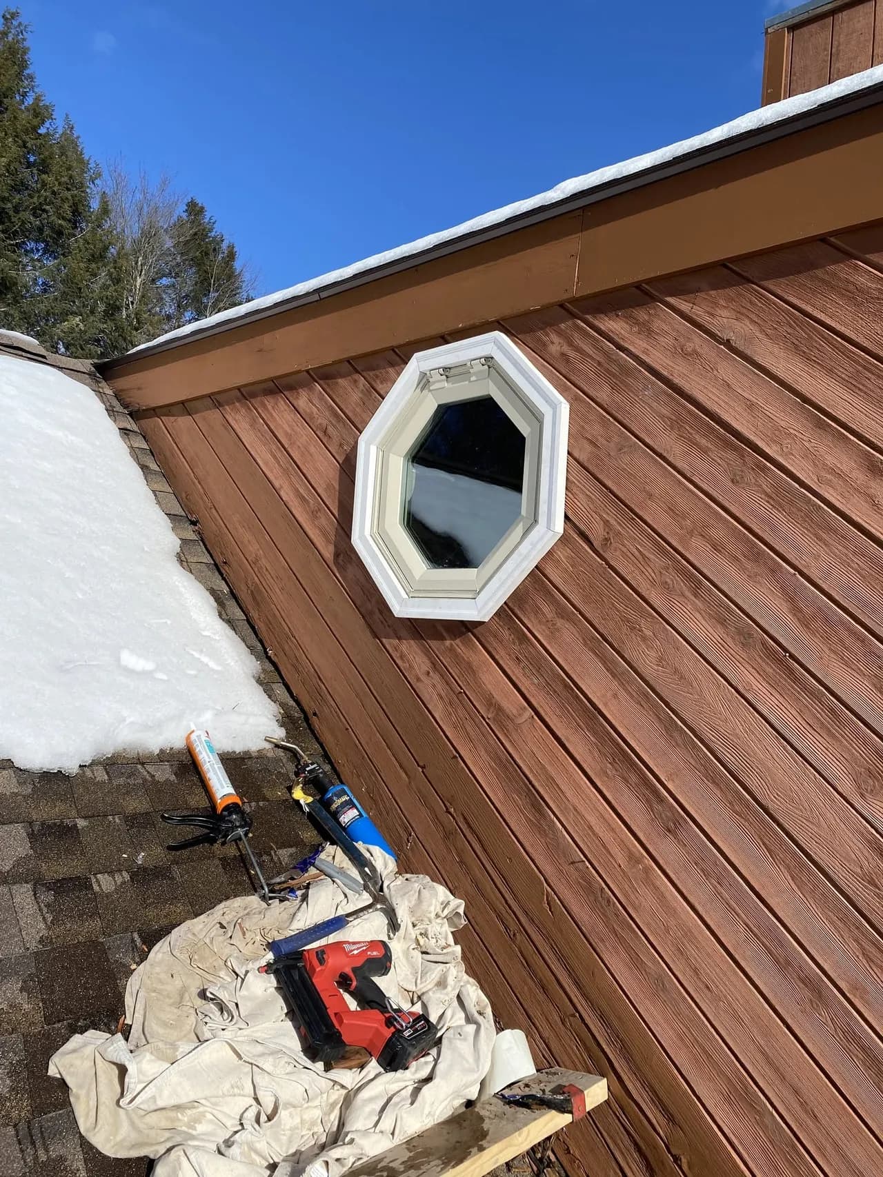 Octagonal window newly installed in brown wood siding above a snow-covered roof with tools laid out