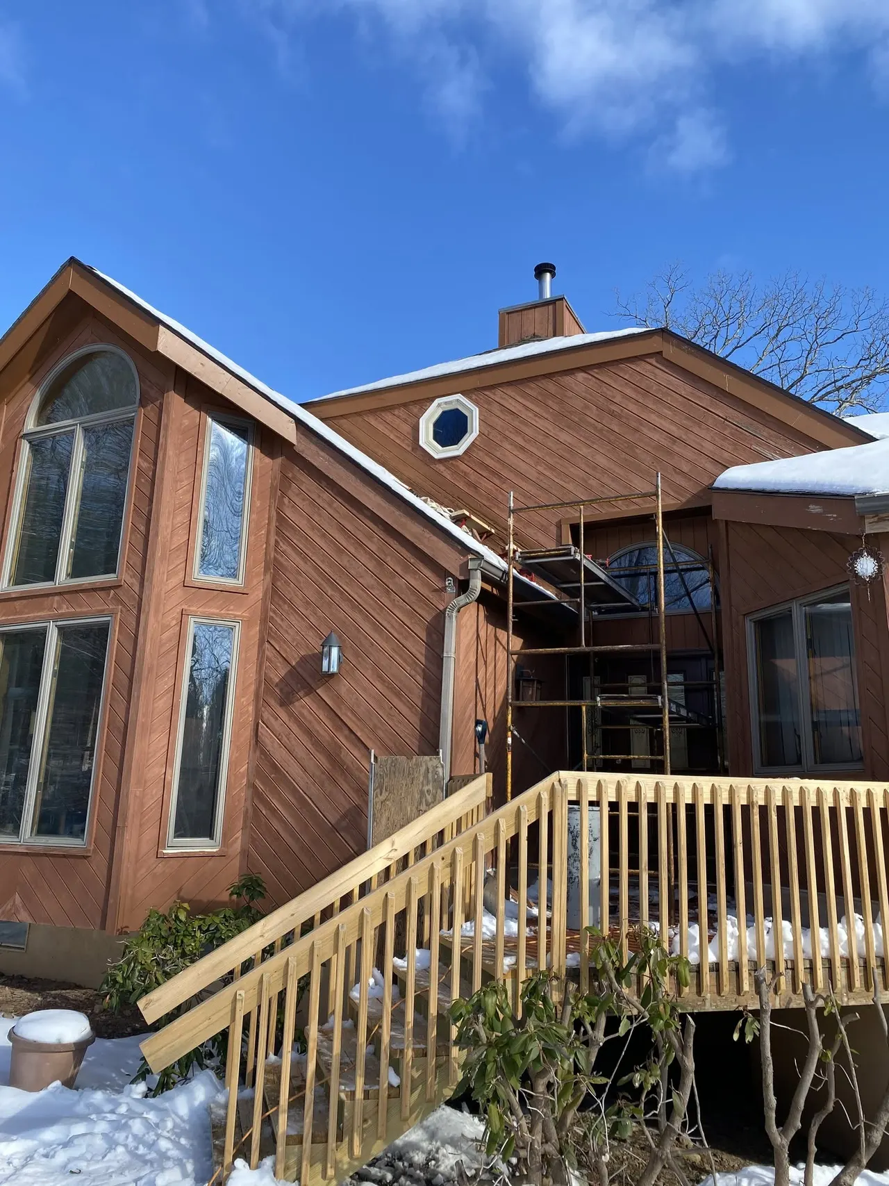 Wood-sided home exterior with tall arched window units and scaffolding beside a new deck in snow